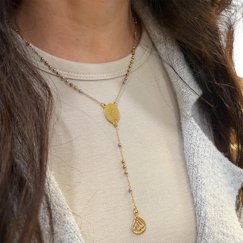 May include: Gold-coloured necklace with a drop pendant. The necklace features small beads and a central oval-shaped pendant. The drop pendant has a teardrop shape with an engraved design. The necklace is worn on a person.