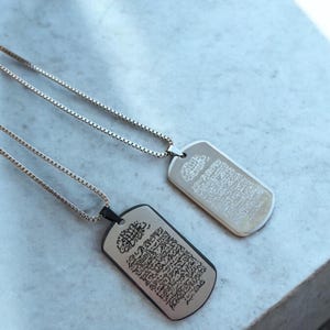 Puede incluir: Dos collares tipo placa de identificación con escritura intrincada. Una placa es plateada con una cadena plateada, la otra es negra con una cadena plateada. Las placas son rectangulares con esquinas redondeadas.