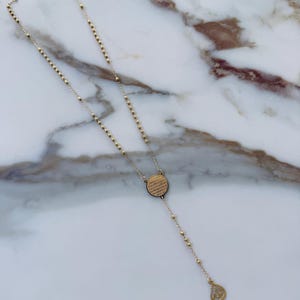 May include: Gold rosary necklace with a circular pendant with etched text and a teardrop charm. The necklace features small, round beads and a delicate chain, set against a marble background.