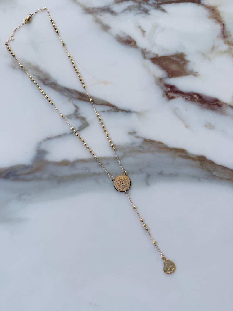 May include: Gold rosary necklace with a circular pendant with etched text and a teardrop charm. The necklace features small, round beads and a delicate chain, set against a marble background.
