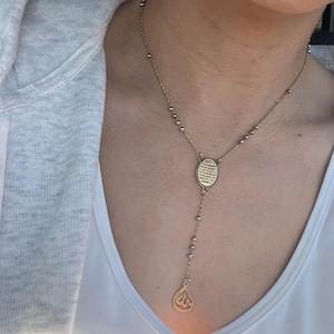 May include: Gold-coloured necklace with an oval pendant and a teardrop charm. The necklace features a delicate chain with small gold and silver beads. The pendant has an engraved design, and the necklace hangs down the chest.