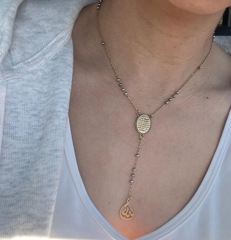May include: Gold-coloured necklace with an oval pendant and a teardrop charm. The necklace features a delicate chain with small gold and silver beads. The pendant has an engraved design, and the necklace hangs down the chest.