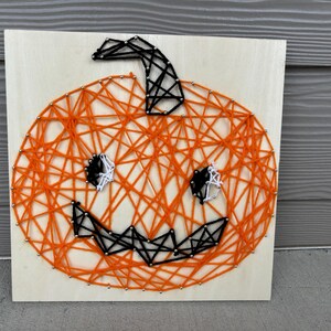 May include: Orange and black string art pumpkin with a black stem and white eyes. The pumpkin is on a wooden background.