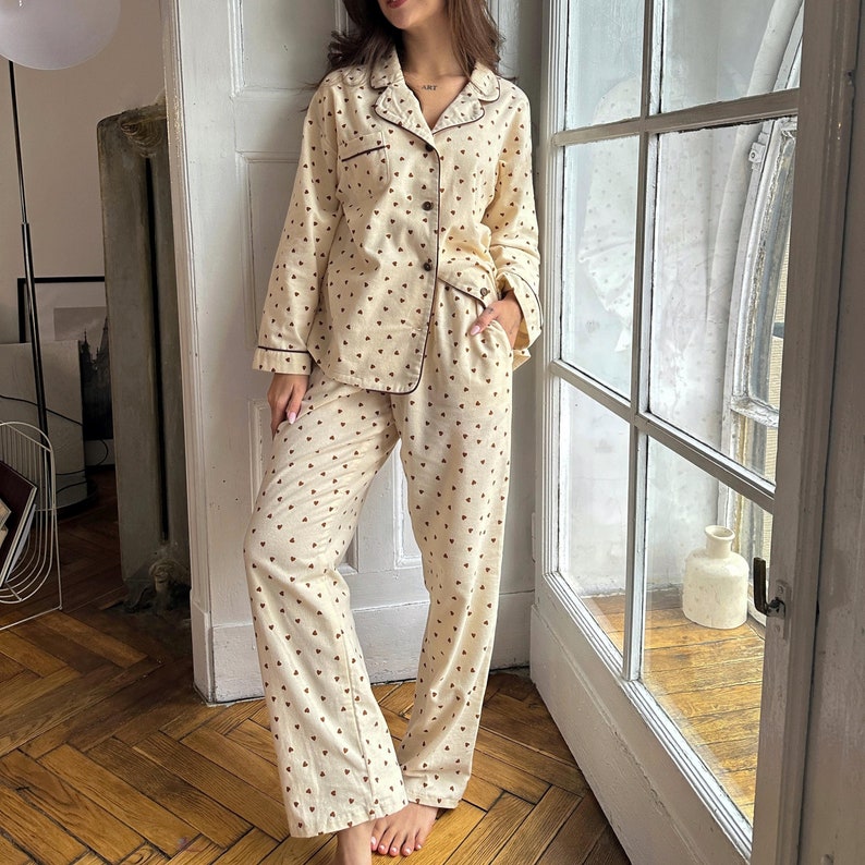 May include: Cream-colored pajama set with a heart pattern. The long-sleeved top has a collared neckline and button-down front. The pants are loose-fitting. The set is trimmed with a dark brown piping.