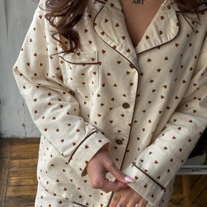 May include: Cream-colored pajama set with a brown heart pattern. The set includes a button-down shirt with a chest pocket and matching pants. Brown trim accents the collar, cuffs, and pocket. The word "ART" is tattooed on the chest.