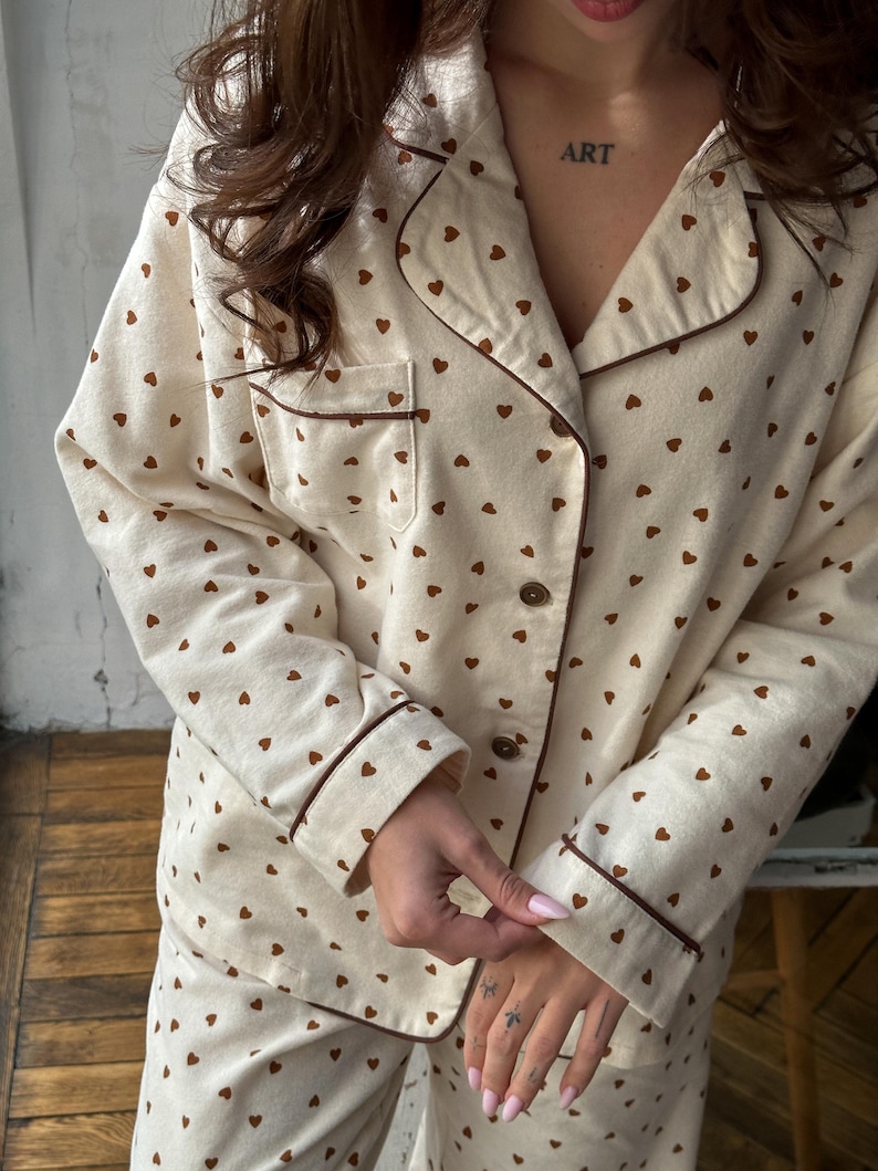 May include: Cream-colored pajama set with a brown heart pattern. The set includes a button-down shirt with a chest pocket and matching pants. Brown trim accents the collar, cuffs, and pocket. The word "ART" is tattooed on the chest.