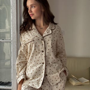 May include: Cream-colored pajama set with a brown heart pattern. The long-sleeved button-down shirt and matching pants have brown trim. The word "ART" is tattooed on the person's neck.