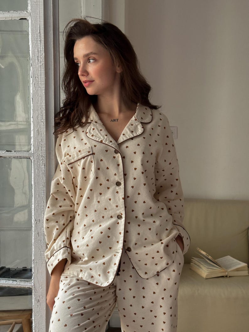 May include: Cream-colored pajama set with a brown heart pattern. The long-sleeved button-down shirt and matching pants have brown trim. The word "ART" is tattooed on the person's neck.