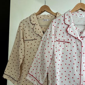 May include: Two sets of pajamas hanging on wooden hangers. One set is cream-colored with brown heart patterns and trim. The other set is white with red heart patterns and trim. Both sets have a button-down top and matching bottoms.