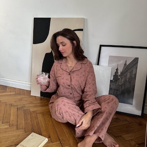 May include: A woman wearing a pink floral pajama set with long sleeves and pants. She is sitting on a wooden floor with a glass of water in her hand. There is a book titled "Fahrenheit 451" on the floor next to her.