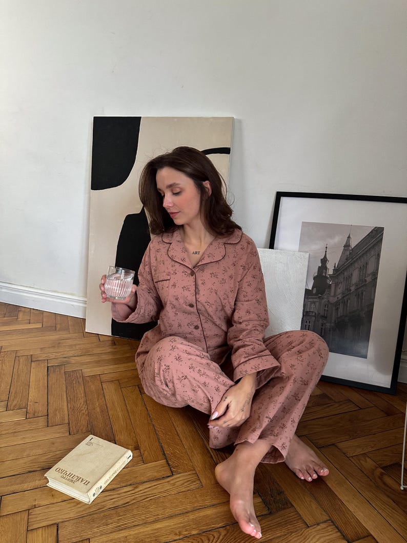 May include: A woman wearing a pink floral pajama set with long sleeves and pants. She is sitting on a wooden floor with a glass of water in her hand. There is a book titled "Fahrenheit 451" on the floor next to her.