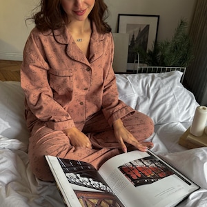 May include: A woman wearing a pink floral pajama set with a button-down top and long pants. She is sitting on a white bed and reading a book with a black cover. The book is open to a page with a picture of a wooden door.