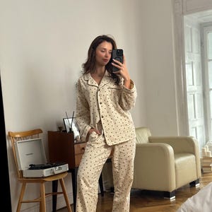 May include: Cream-colored pajama set with a repeating pattern of small, dark brown dots. The set includes a button-down shirt with a notched collar and long sleeves, and matching straight-leg pants. The pajamas have dark brown trim.