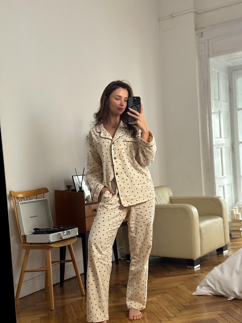 May include: Cream-colored pajama set with a repeating pattern of small, dark brown dots. The set includes a button-down shirt with a notched collar and long sleeves, and matching straight-leg pants. The pajamas have dark brown trim.