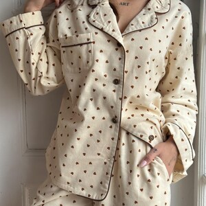 May include: Cream-colored pajama set with a repeating pattern of small brown hearts. The long-sleeved top has a button-down front, a breast pocket, and brown trim. The matching pants have a relaxed fit. The word "ART" is visible on the person's neck.