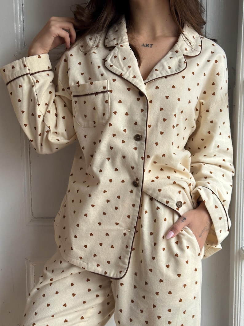 May include: Cream-colored pajama set with a repeating pattern of small brown hearts. The long-sleeved top has a button-down front, a breast pocket, and brown trim. The matching pants have a relaxed fit. The word "ART" is visible on the person's neck.