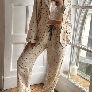 May include: Cream-colored pajama set with a heart pattern. The set includes a button-down top with black trim and matching pants with a drawstring waist. The pajamas are made of a soft fabric and are suitable for lounging or sleeping.