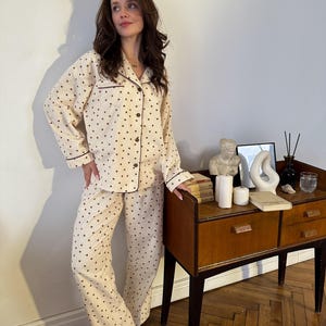 May include: Cream-colored pajama set with a heart pattern. The long-sleeved top and pants are adorned with small, dark brown hearts. The set features dark brown trim and buttons. The pajamas are made of a soft, comfortable material.