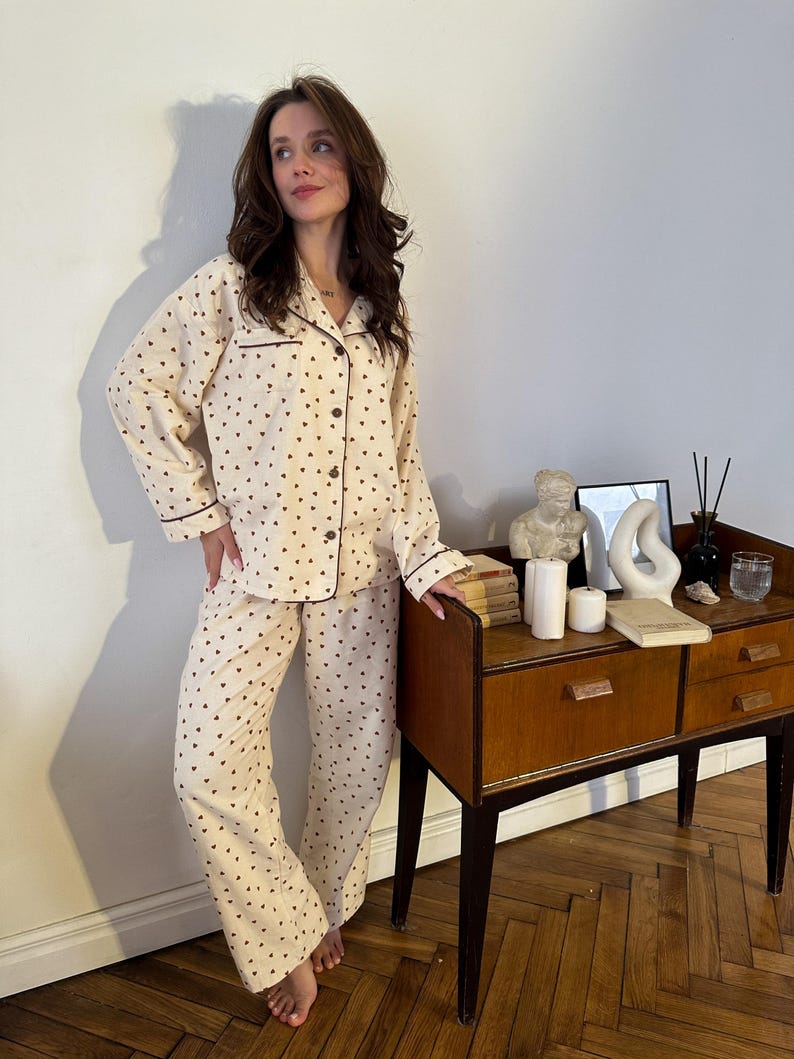 May include: Cream-colored pajama set with a heart pattern. The long-sleeved top and pants are adorned with small, dark brown hearts. The set features dark brown trim and buttons. The pajamas are made of a soft, comfortable material.