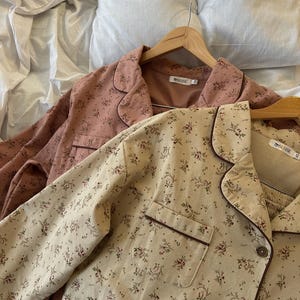 May include: Two sets of floral-patterned pajamas on wooden hangers. One set is a light pink with dark brown trim, and the other is beige with dark brown trim. The pajamas have a button-down top and a pocket on the chest.