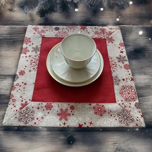 以下が含まれることがあります： 赤色のセンターと赤と白の雪の結晶のボーダーが付いたお祝いのクリスマスプレースマット。 金色のトリムが付いた白いティーカップとソーサーのセットがプレースマットの上に置かれ、お祝いのテーブルセッティングを作成します。