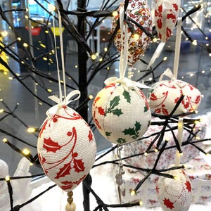 Puede incluir: Primer plano de un árbol decorativo negro adornado con varios adornos navideños de tela. Los adornos presentan patrones de acebo y florales rojos y verdes, y están suspendidos por cintas blancas. Algunos adornos tienen cuentas colgantes.