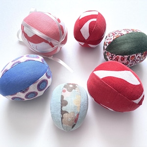 May include: A collection of six fabric Easter eggs in various colors and patterns. The eggs feature designs in red, blue, green, and floral prints. Some eggs have white ribbon accents.