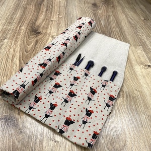 May include: A beige and brown fabric makeup brush roll with a black and white pattern of cartoon dogs wearing red berets. The roll has five slots for brushes and is rolled up.
