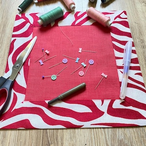 May include: A red sewing mat with a white and red zebra print border. The mat holds sewing pins, scissors, a pen, and spools of thread in various colors. A craft-related item.