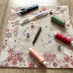 May include: A collection of sewing supplies on a decorative fabric square. Includes spools of thread in pink, green, and red, along with pins, buttons, scissors, and a marking pen. The fabric has a snowflake pattern.