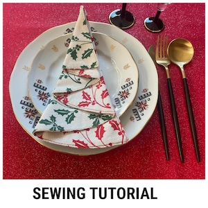 May include: A white and red Christmas tree napkin folded into a festive shape. The napkin is decorated with green holly leaves and red berries. The napkin is placed on a white plate with a gold rim. The plate is on a red tablecloth.