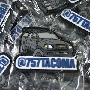 May include: A close-up of a black and white illustration of a Toyota Tacoma pickup truck with a roof rack. The truck has a blue and white sticker that says "@757TACOMA".