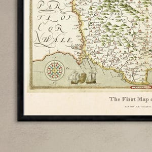 May include: Framed antique map of Devon, England, with a black frame. The map displays detailed cartography, including illustrations of ships and a compass rose. The text at the bottom reads "The First Map of Devon" by Saxton, Christopher, 1575.