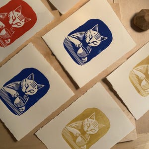 K&ouml;nnte beinhalten: Vier Karten mit wei&szlig;em Hintergrund und einer Katzenillustration in verschiedenen Farben. Die Katze trinkt aus einer Sch&uuml;ssel. Die Farben sind rot, blau, gelb und wei&szlig;.