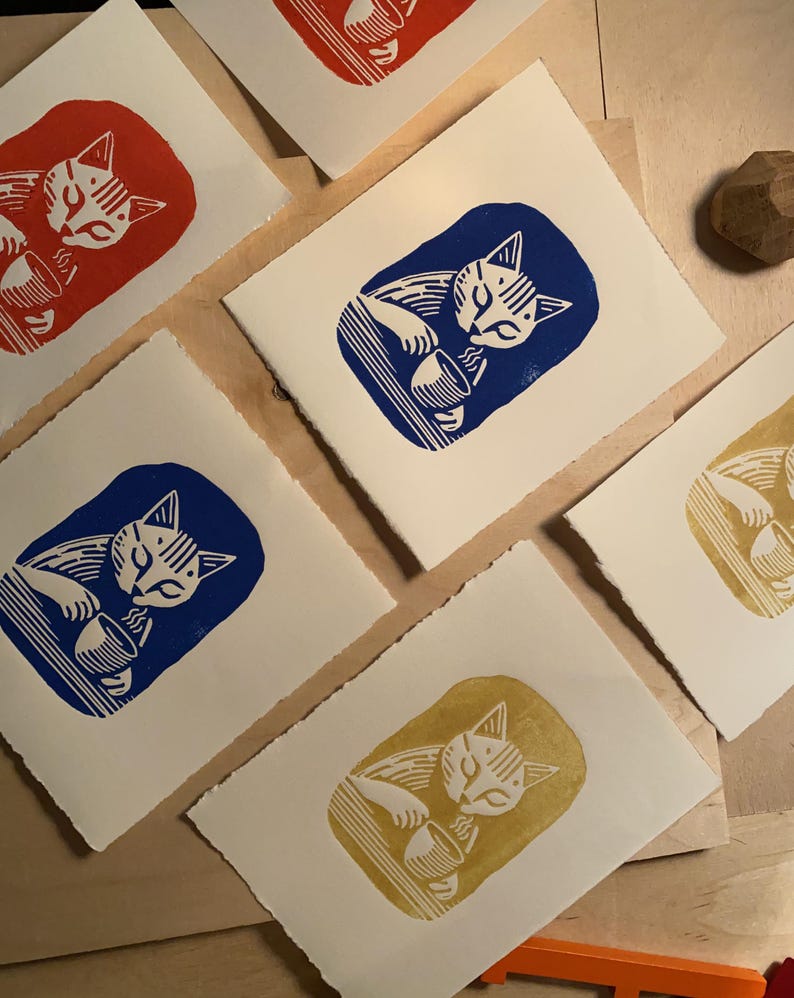 K&ouml;nnte beinhalten: Vier Karten mit wei&szlig;em Hintergrund und einer Katzenillustration in verschiedenen Farben. Die Katze trinkt aus einer Sch&uuml;ssel. Die Farben sind rot, blau, gelb und wei&szlig;.
