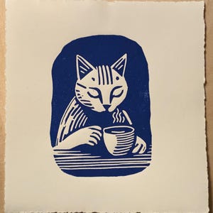 K&ouml;nnte beinhalten: Ein blauer und wei&szlig;er Linolschnitt eines Katers, der aus einer Tasse trinkt. Der Kater sitzt an einem Tisch und h&auml;lt eine dampfende Tasse in seinen Pfoten.