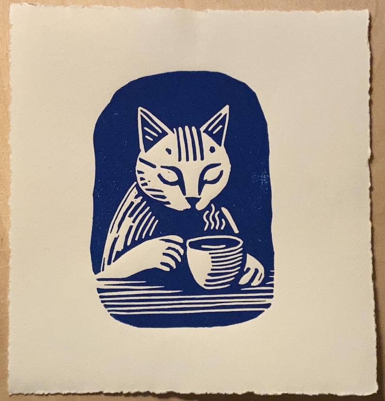 K&ouml;nnte beinhalten: Ein blauer und wei&szlig;er Linolschnitt eines Katers, der aus einer Tasse trinkt. Der Kater sitzt an einem Tisch und h&auml;lt eine dampfende Tasse in seinen Pfoten.
