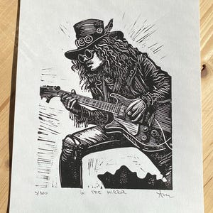 Peut inclure: Gravure sur bois en noir et blanc d'un musicien jouant de la guitare. Le musicien porte un chapeau et une veste en cuir. L'estampe est intitulée "Dans le miroir" et numérotée 5/300.