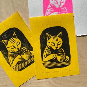 K&ouml;nnte beinhalten: Zwei gelbe Karten mit schwarzen Linolschnitten von einer Katze, die Kaffee trinkt. Der Text "COFFEE LINOCUT" ist auf der Unterseite der Karte rechts gedruckt.