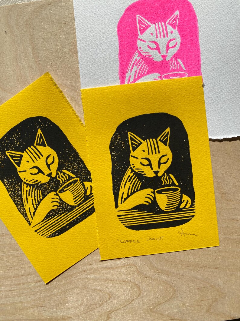 K&ouml;nnte beinhalten: Zwei gelbe Karten mit schwarzen Linolschnitten von einer Katze, die Kaffee trinkt. Der Text "COFFEE LINOCUT" ist auf der Unterseite der Karte rechts gedruckt.