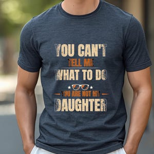 Puede incluir: Una camiseta de manga corta de color azul oscuro jaspeado con el texto desgastado en beige y naranja "YOU CAN'T TELL ME WHAT TO DO YOU ARE NOT MY DAUGHTER" y un gráfico de gafas de sol con montura naranja centrado entre las líneas.