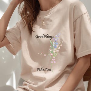 Op de afbeelding: Beige T-shirt met de tekst "Good Things Take Time" en een bloemenontwerp in blauw, paars en roze. Het shirt heeft korte mouwen en een ronde hals. Een gouden ketting is zichtbaar.