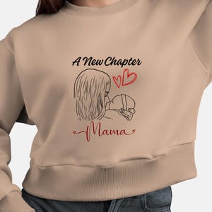 Puede incluir: Sudadera beige con un gráfico negro y rojo. El gráfico presenta un dibujo lineal de una madre y un bebé, con las palabras "A New Chapter" y "Mama" en escritura negra y roja, junto con dos corazones rojos.