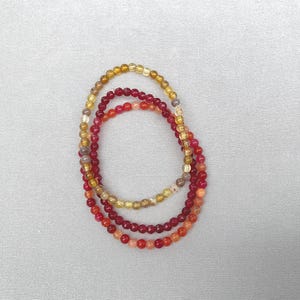 May include: Three circular beaded bracelets arranged in a nested formation. One bracelet features translucent yellow and amber-toned beads, while the other two consist of small, polished spherical beads in varying shades of deep red, crimson, and orange.