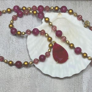 May include: A necklace featuring a teardrop-shaped, deep red pendant and a strand of alternating gold and magenta beads. The necklace is displayed on a white seashell against a textured gray background.