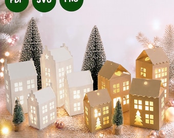 May include: A collection of white and gold paper house luminaries with cut-out windows and doors, illuminated from within. The set includes various architectural styles, accompanied by miniature bottle brush trees and green circular icons labeled PDF, SVG, and PNG.