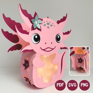 Puede incluir: Una linterna de papel con forma de ajolote rosa, branquias de color rosa oscuro, una flor de purpurina plateada y un centro iluminado. El diseño incluye recortes florales en los laterales e iconos de formato PDF, SVG y PNG.