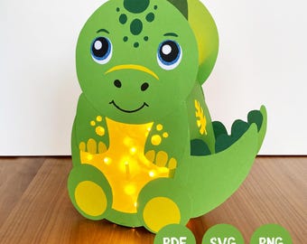 Könnte beinhalten: Eine grüne Papier-Dinosaurier-Laterne mit großem Kopf, blauen Augen, lächelndem Mund und einem gelben Bauch, der von innen warm beleuchtet wird. Der Dinosaurier hat einen geschwungenen Schwanz und gelbe Füße, mit den Beschriftungen PDF, SVG und PNG.
