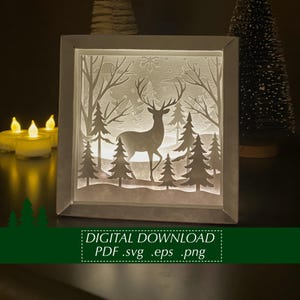 Könnte beinhalten: Eine beleuchtete, gerahmte Papierschnitt-Szene mit einem Hirsch in einem Wald mit Bäumen und Schneeflocken. Der Rahmen ist weiß, und die Szene wird von innen beleuchtet. Der Text unten lautet "DIGITAL DOWNLOAD PDF .svg .eps .png".