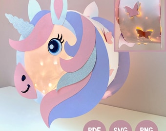 Könnte beinhalten: Eine zylindrische Papierlaterne in Form eines Einhornkopfes mit einem silbernen Glitzerhorn, einer rosa und violetten Papiermähne und einem großen blauen Auge. Die Laterne wird von innen beleuchtet und strahlt durch Schmetterlingsausschnitte.